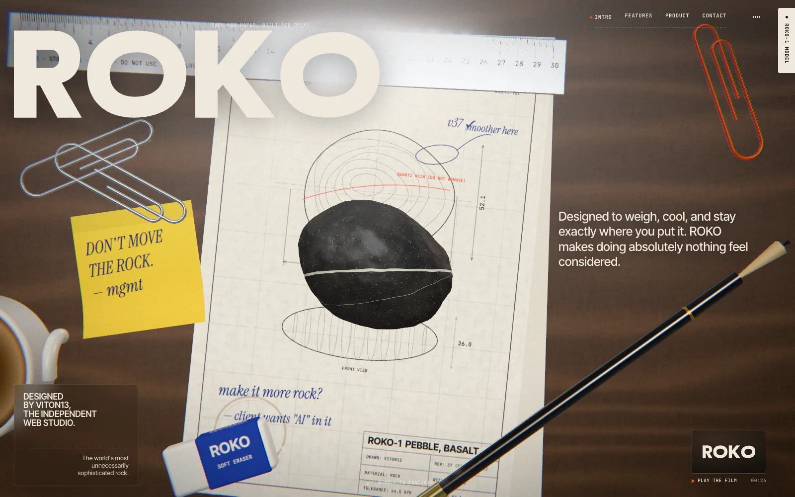The ROKO home page: a top-down flat-lay on a walnut desk — a technical drawing of the pebble under a real-time 3D basalt stone with a white vein, a pencil, a steel ruler, paperclips, a coffee cup and a yellow sticky note reading “Don’t move the rock. — mgmt”, with the huge ROKO wordmark at the top left.